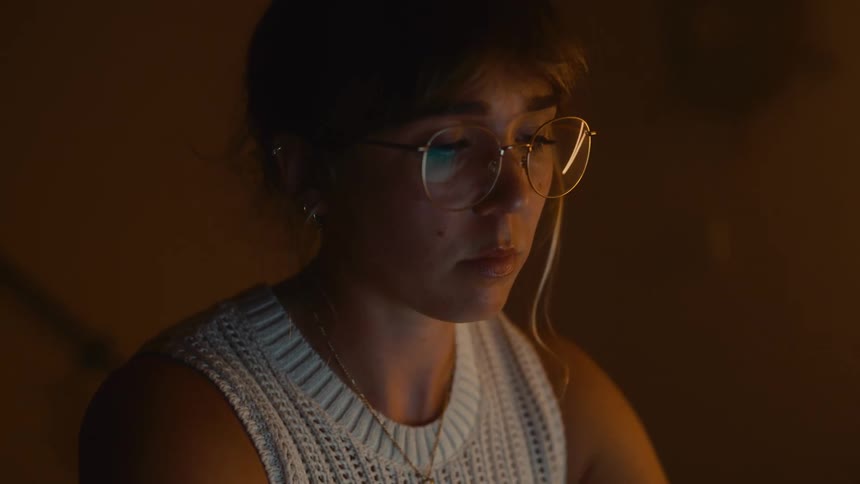 A close view of a woman wearing glasses and a white sleeveless top looks down bathed in the dim warm light of a dark room