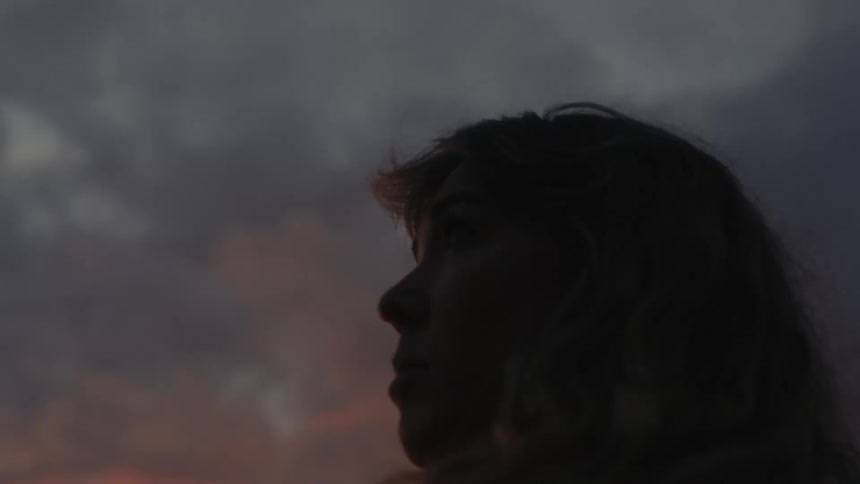 A young woman with long hair standing outdoors beneath a cloudy sunset sky
