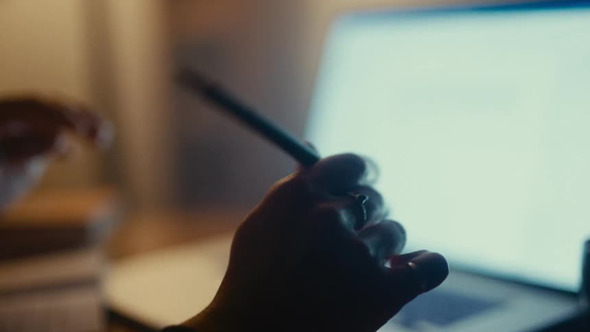 Hand holds a pen in front of a lit screen in a dimly lit setting