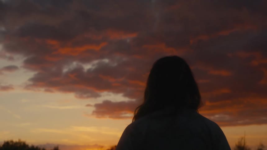 Woman stands near a forest looking at the red sky while the sun sits on the horizon