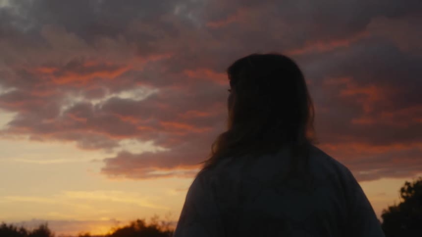 Woman stands outdoors under a red tinted sky and looks around in an area near the woods