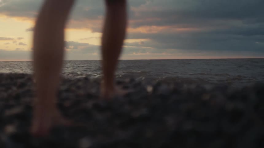 Legs of a person walking barefoot on a gravel strip near the waves