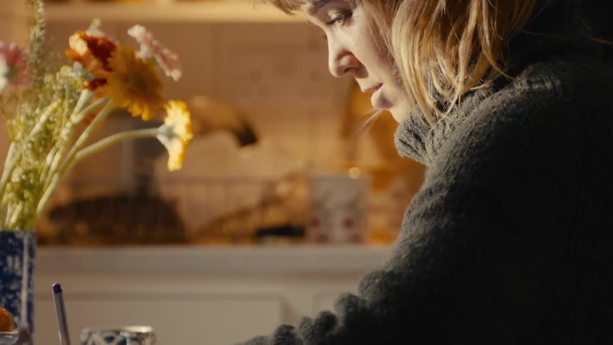 A woman in a cozy knit sweater leans forward in concentration her profile illuminated by warm light as she sits at a table with a vase of flowers and writes something