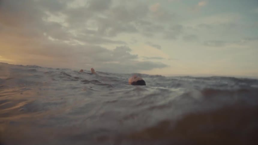 A lowangle shot captures a woman struggling in dark choppy ocean water as she attempts to keep her head above the rolling waves against a dramatic cloudy sky illuminated by the sunset