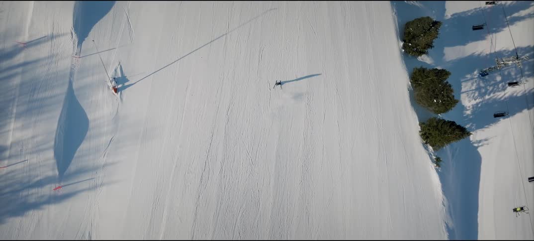 A person skiing down a snowy mountain trail at midday while others ascend on a ski lift to the right