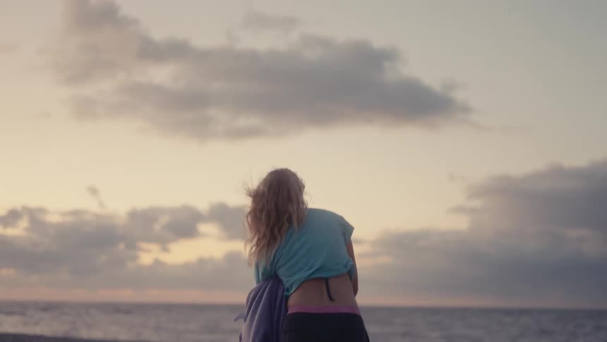 Young woman stands on the beach looking at the water as she takes off her shirt