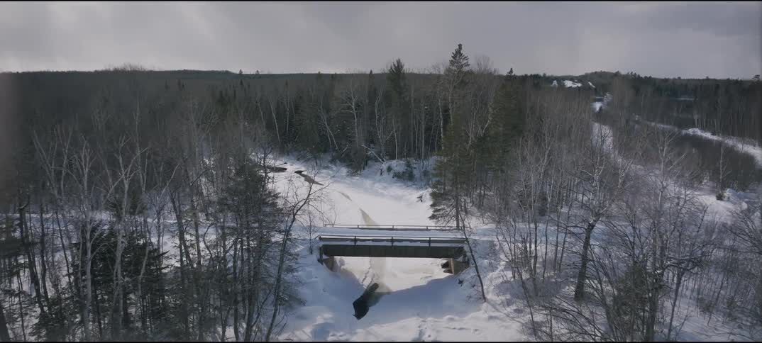 A drone shot captures a winter landscape showing a bridge spanning a partially frozen or snowcovered creekravine surrounded by a dense forest the camera descends and tilts down eventually showing two snowmobiles driving across the bridgethe scene