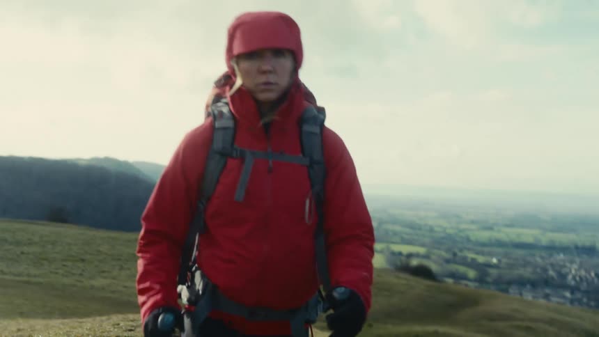 A hiker in a red jacket treks along a grassy ridge overlooking a vast valley