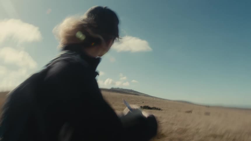 Young adult woman wearing a baseball cap writing down notes on a notepad and looking at a natural landscape outdoors during day time