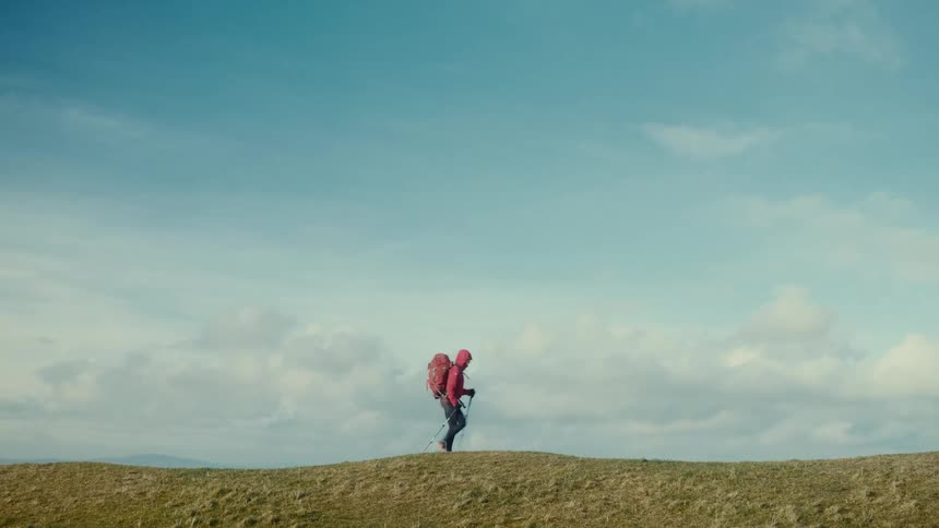 Person with red backpack walks across grassy hill under partly cloudy sky