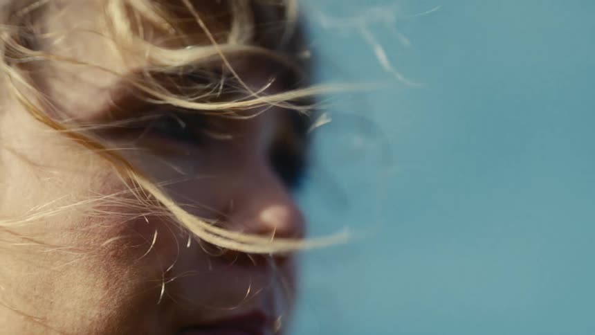 A closeup shot of a womans profile outdoors with her blonde hair strands gently blowing across her face due to the wind against a bright hazy blue background