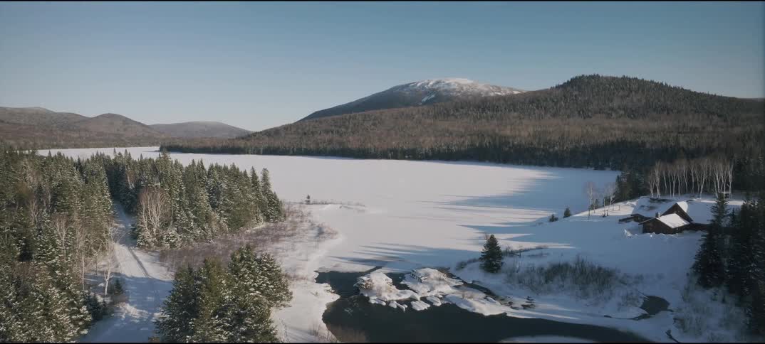 An aerial shot overlooks a snowy winter landscape featuring a frozen lake pine trees a cabin and a lone person walking on a path