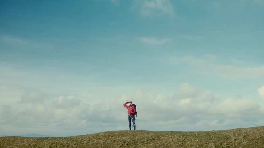 Female person wearing a red hood and carrying a backpack looking around from the top of a slope outdoors