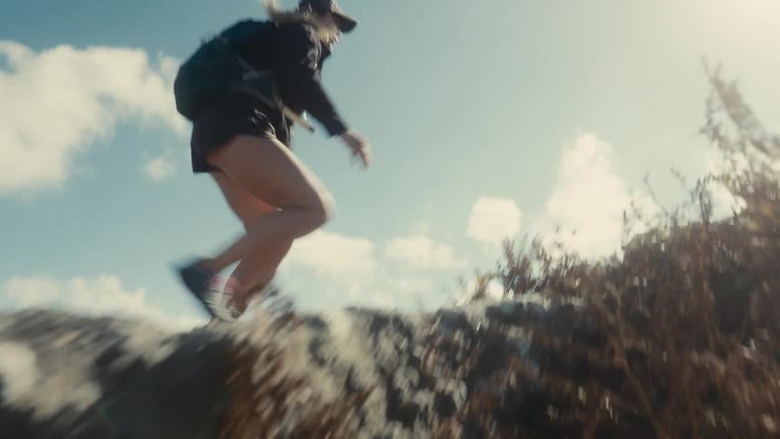 A fit woman wearing shorts a dark jacket and a backpack jumps powerfully over a boulder on a rocky scrub covered hill against a bright sky