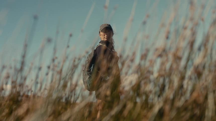 A female hiker with a backpack stands in a field framed by tall blurred grass in the foreground