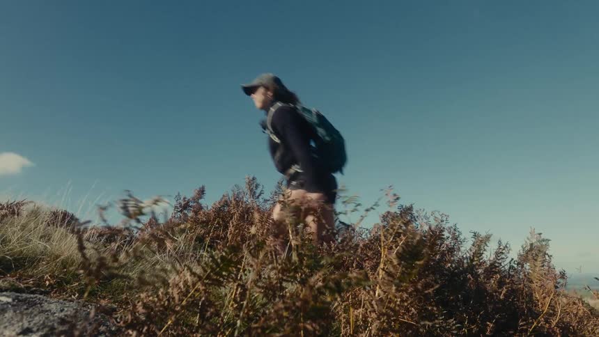 A person wearing a cap and backpack hikes uphill through scrub and brush moving from the right side of the frame towards the center the backdrop is a clear blue sky suggesting an outdoor adventure or trek