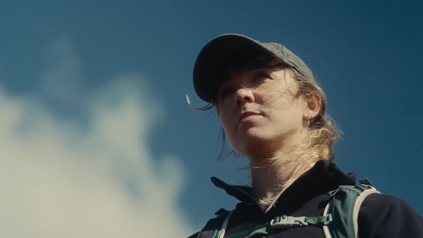 A female hiker looks toward the sky with a thoughtful expression her face warmed by sunlight as the wind gently blows strands of hair across her face