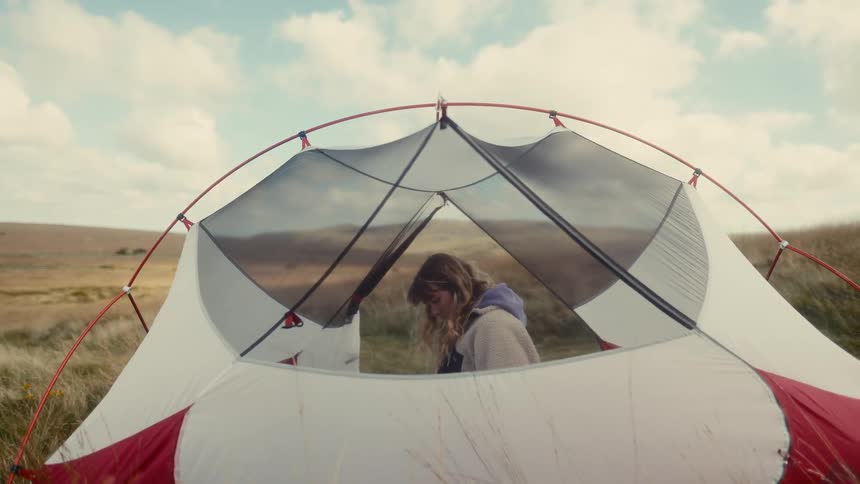 A woman wearing a pale purple hoodie and a white fleece jacket looks down while sitting inside a tent pitched in a grassy field under a cloudy sky