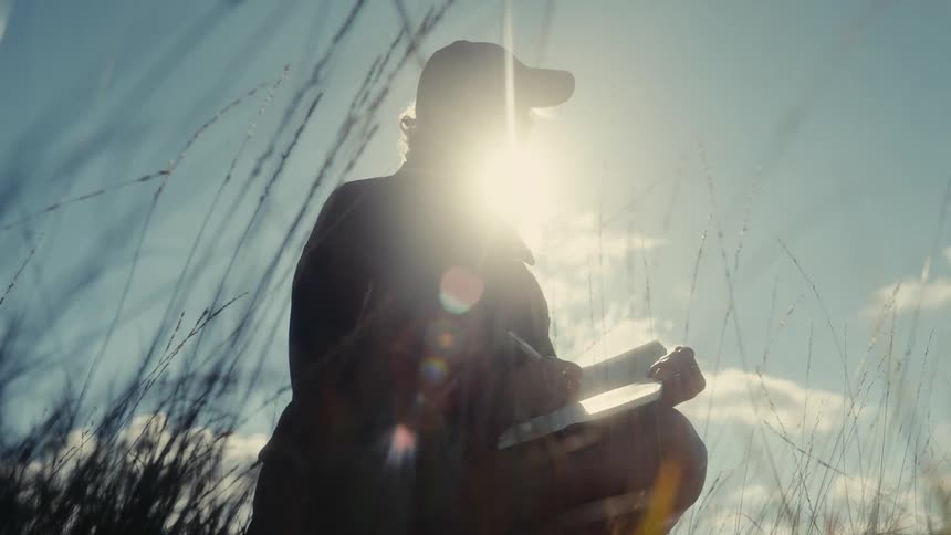 Person writing in a notebook while crouching among tall grass