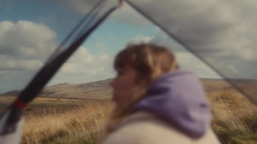 Woman in a tent set in a grassland on a cloudy day looks around