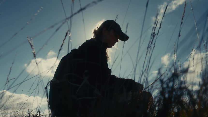 Person sitting among tall grass appears in silhouette against the bright sky