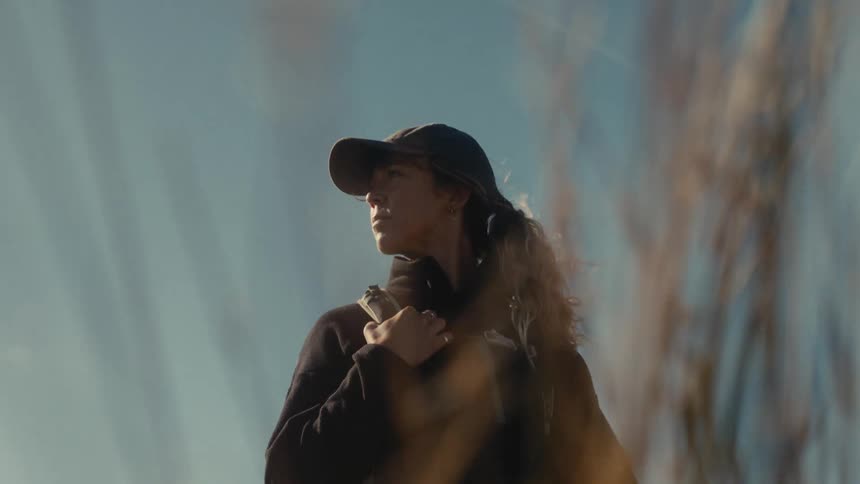 A determined woman in a dark hoodie wearing a backpack is looking forward amongst golden reeds