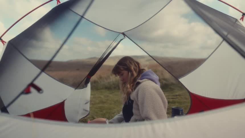 From the cozy interior of a tent a woman sits outside in a vast open field writing thoughtfully in a journal under a partly cloudy sky