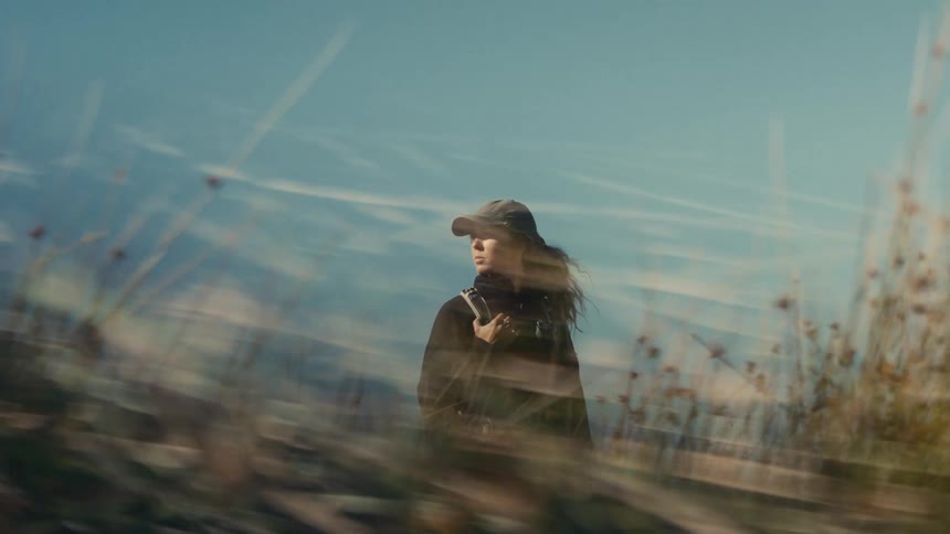A young woman in a cap stands in a field framed by tall blurred grass in the foreground