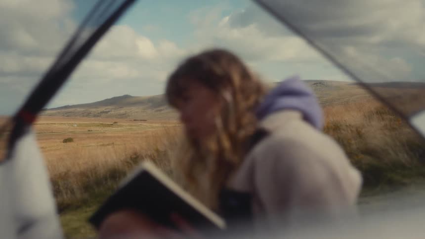 A young blonde woman is in a tent on an outlying slope with a large grassy field behind her and she is reading