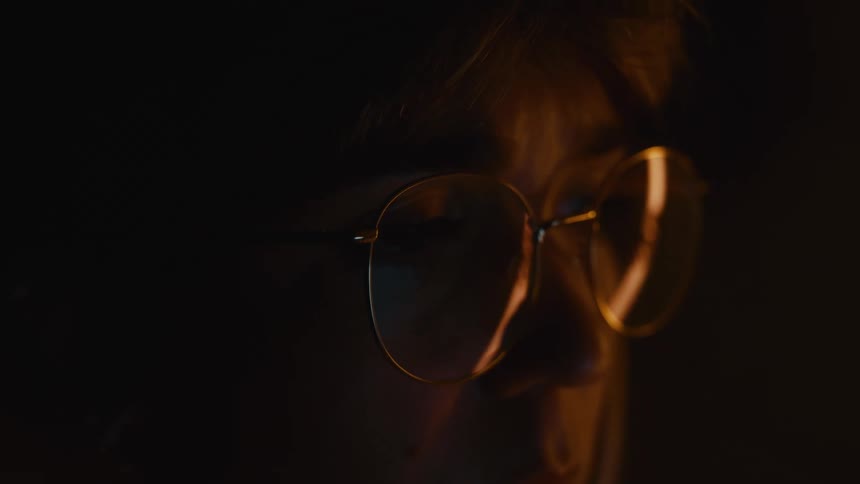 Partially lit face with round eyeglasses reflecting warm light