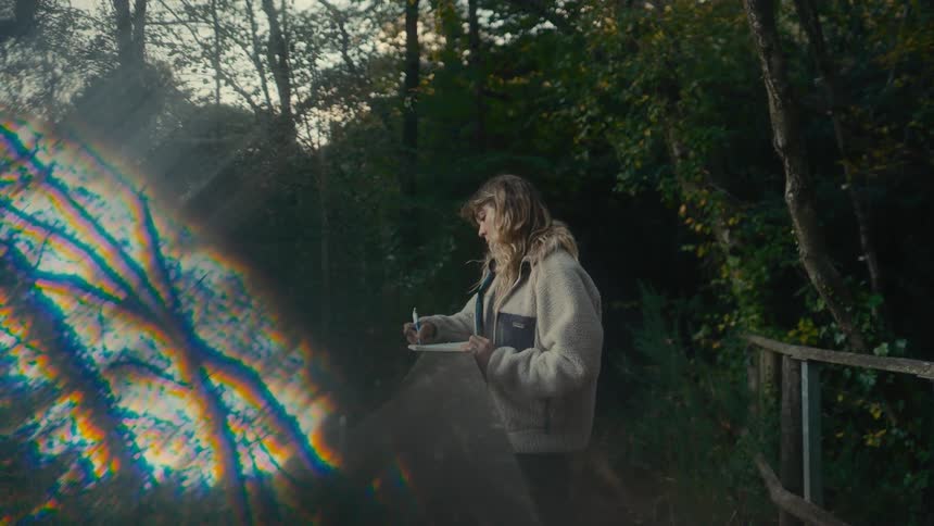 Surrounded by nature on a quiet forest path an artist pauses from sketching in her notebook her thoughtful gaze framed by a vivid prismatic reflection