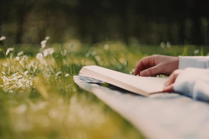 A close view of a womans hands wearing a white shirt resting on a book in a sunlit grassy field with white flowers