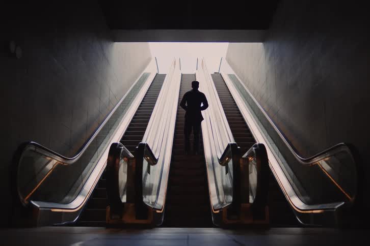 A person is standing on an escalator and going up towards an outside area