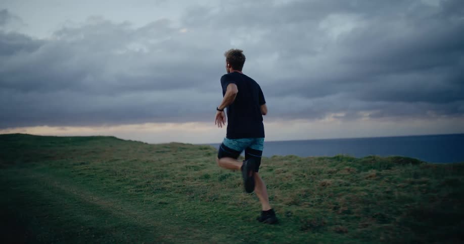 A man runs along a grassy coastal cliff overlooking the ocean under a cloudy sky