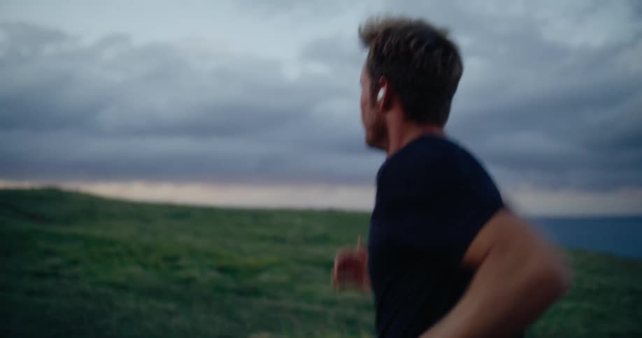 Young adult man with an earpod on his ear running outdoors surrounded by nature during dusk or dawn