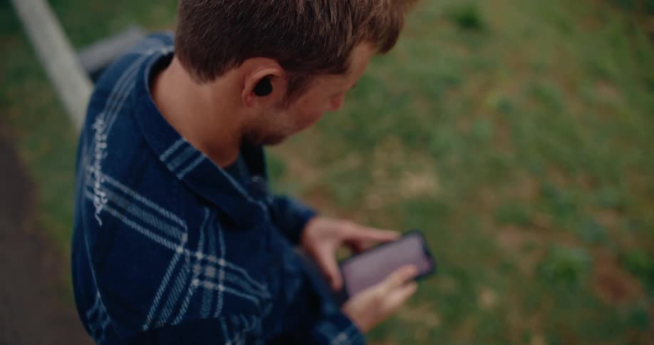 A man with his face turned away is wearing black headphones while fiddling with a cell phone