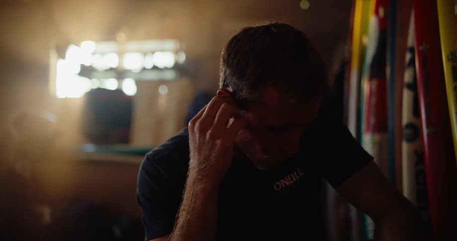 Young adult man sitting on a room with a row of surfboards next to him and putting an earbud inside his ear indoors during day time