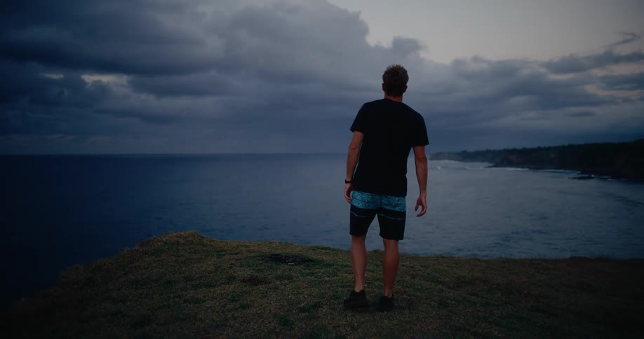 A man wearing a black shirt and patterned shorts stands on a grassy cliff edge overlooking the ocean under a dark dramatic cloudy sky at dusk