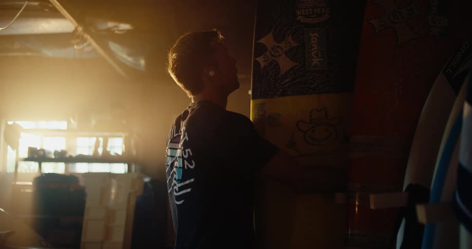 Young man walks over to the wall to pick out a surfboard and carries it under his arm