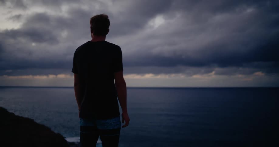 Young adult man standing on a cliff looking at the ocean outdoors during dusk or dawn