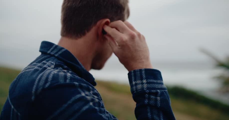 A man in a plaid shirt inserts a wireless earbud while overlooking a coastal landscape
