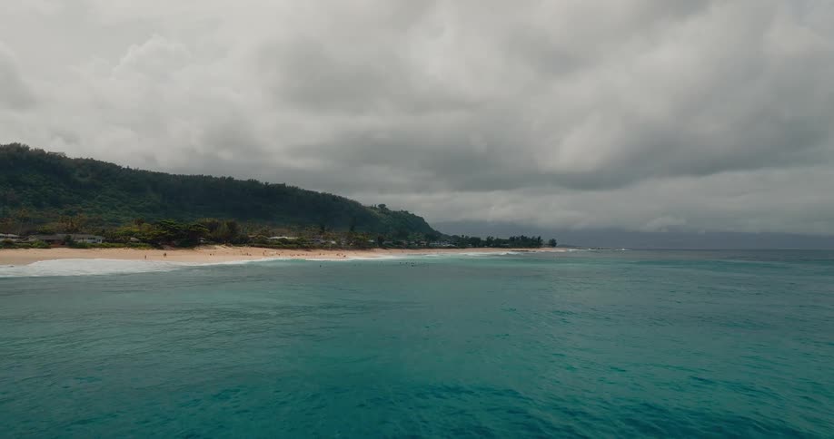 A high angle aerial view captures a wide expanse of turquoise ocean water and a long sandy beach near a dark green hillside under a gray cloudy sky