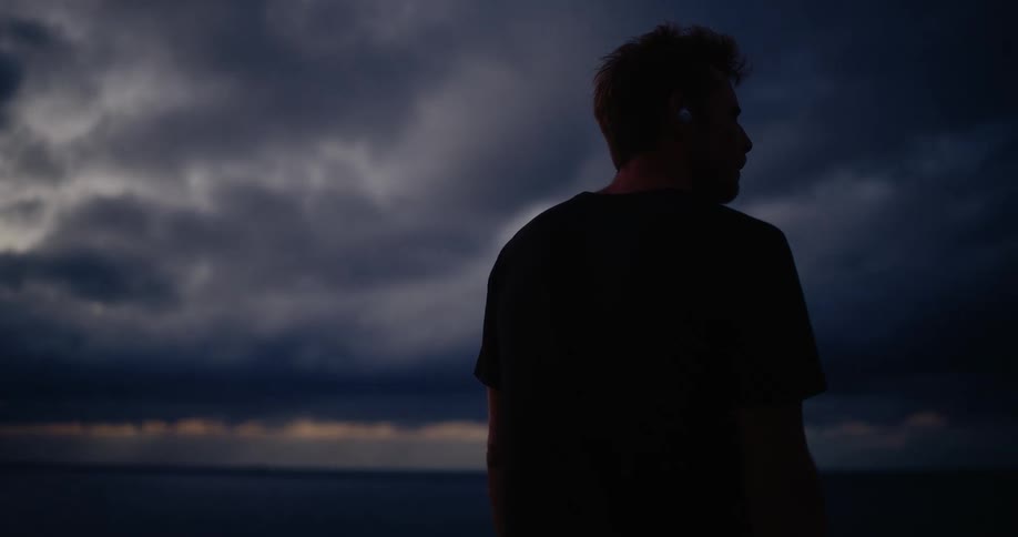 A young man standing on a rocky shoreline watching dark clouds in the evening