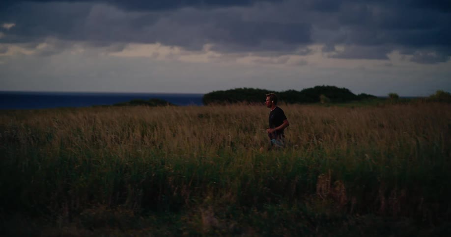 A man wearing a black shirt and dark shorts jogs through tall dry grass on a hilly coastal trail at dusk under a dark cloudy sky
