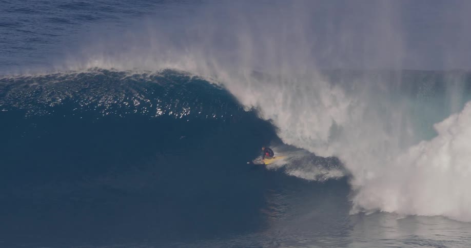 A surfer rides down the face of a colossal breaking wave while safety jet skis float nearby