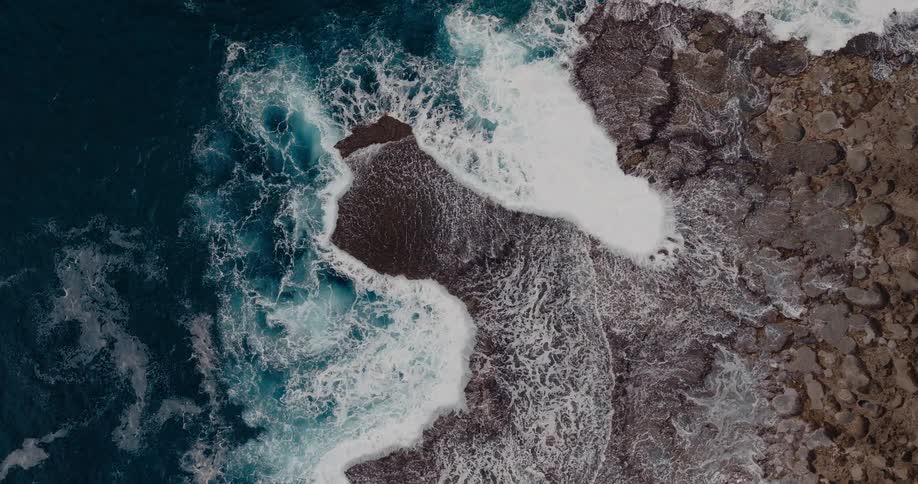 Aerial of clear ocean waves splashing against a rocky coastal area during daytime