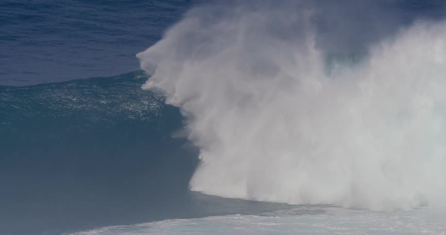 A surfer rides down a massive wave disappearing into the crashing white water before emerging safely near a group of jet skis
