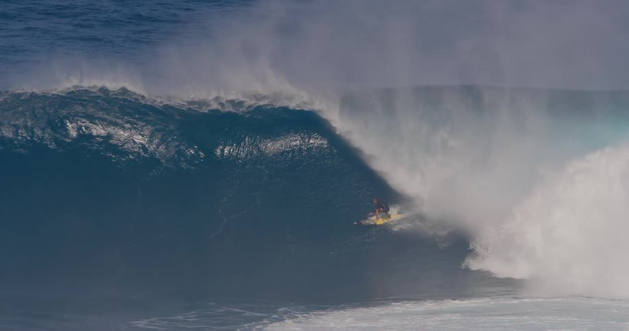A single surfer rides a huge dark blue wave carving along the face as the wave barrels and crashes around them