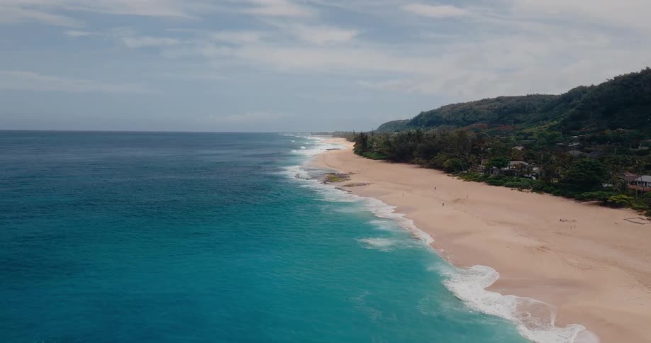 Aerial drone shot shows a long beautiful sandy beach with turquoise ocean water breaking into white waves near a lush green hillside under a partly cloudy sky