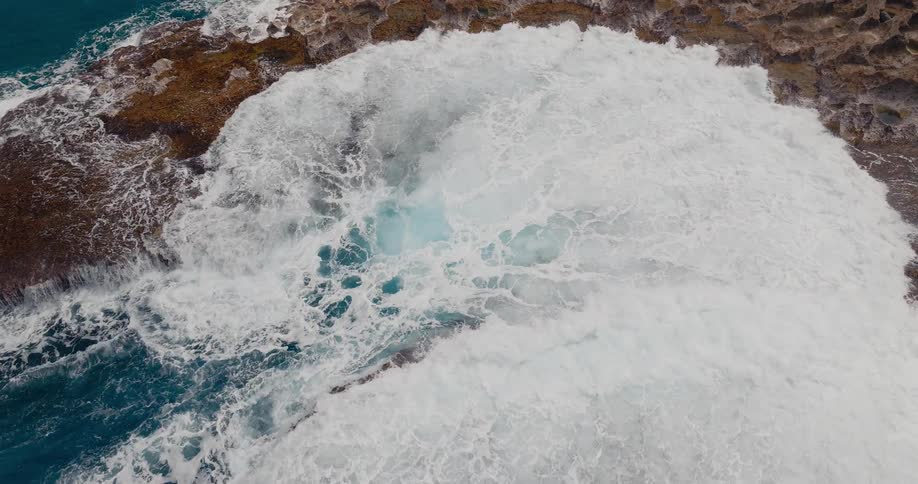 An aerial view shows powerful turquoise ocean waves crashing and churning into a hole or blowhole in a reddish brown rocky shoreline